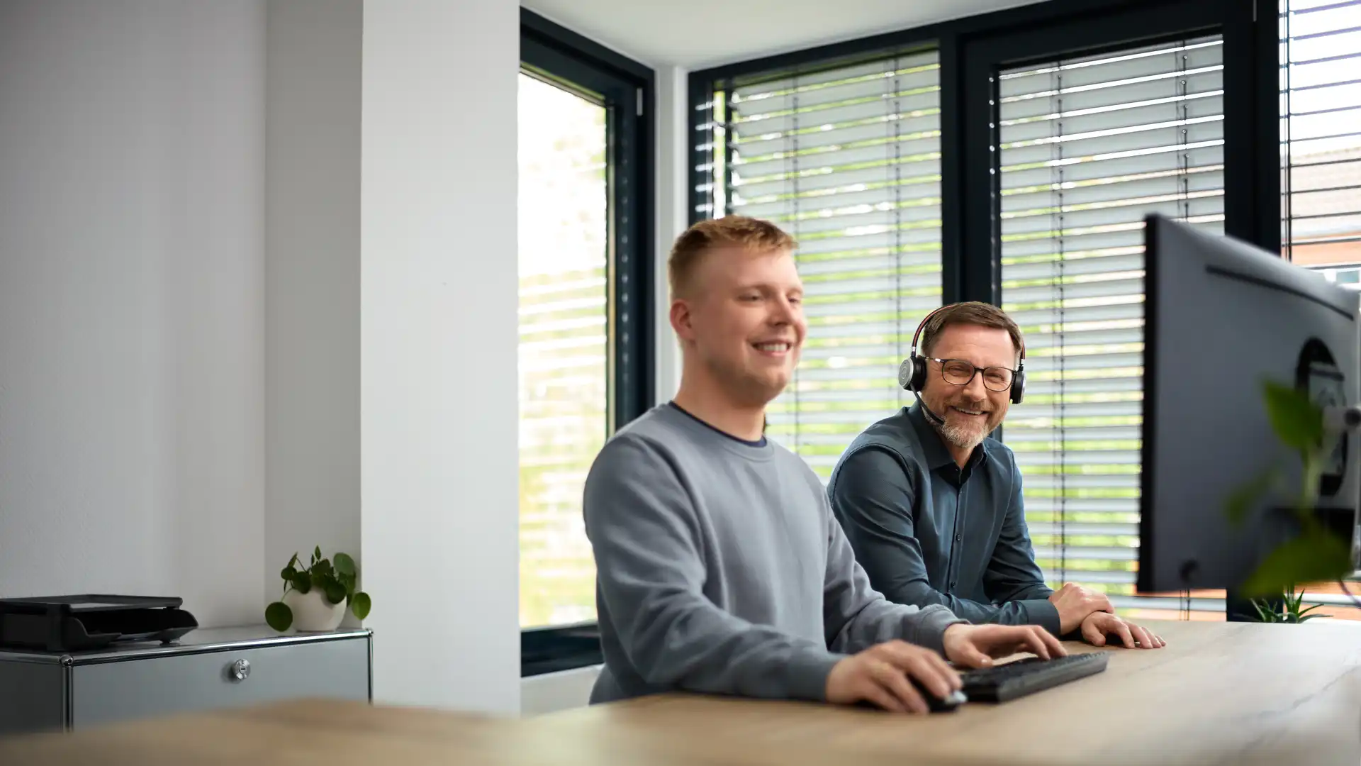Zwei Männer sitzen nebeneinander an einem Schreibtisch, einer von ihnen trägt ein Headset